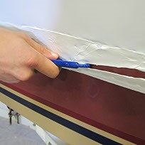 a and holding a tool with a sharp knife end for precision cutting, the sharp end is partially through a cut at the perimeter of a shrink wrap cover on a boat